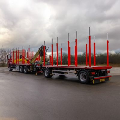 Camion rouge pour le transport de bois avec une remorque, des montants rouges et une grue sur une route humide.